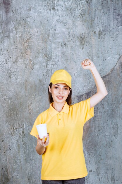 Агент женской службы в желтой форме держит пластиковый стаканчик и показывает знак рукой.
