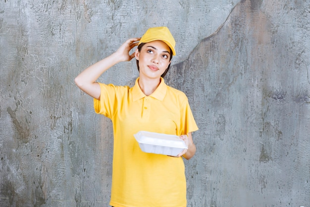 Female service agent in yellow uniform holding a plastic takeaway box and looks confused and thoughtful.