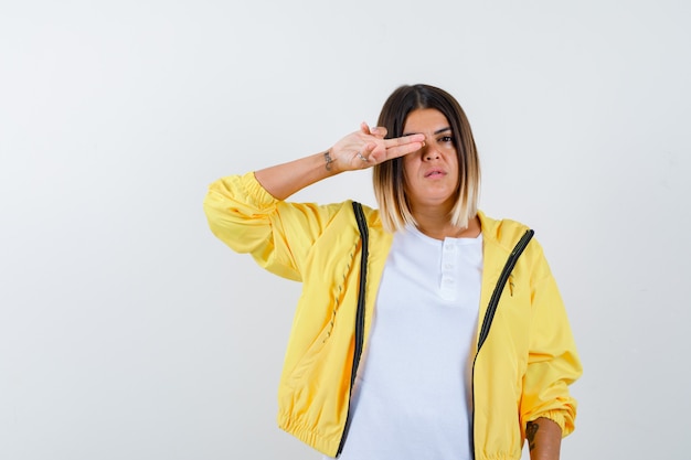 Female showing gun gesture on eye in t-shirt, jacket and looking confident. front view.