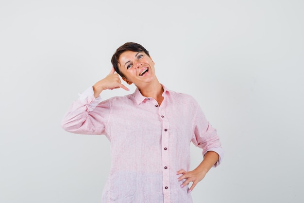 Female showing phone gesture in pink shirt and looking cheerful , front view.