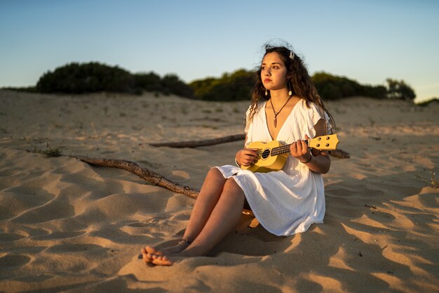 female sitting on a sandy ground while playing a yellow ukulele at the beach