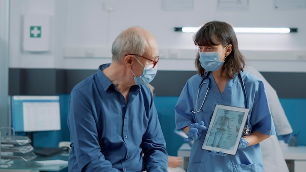 健康診断でフェイスマスクを持った老人にタブレットで人間の骨格のイラストを説明する女性スペシャリスト。骨の検査を示す、オステオパシー診断を備えた看護師保持装置。