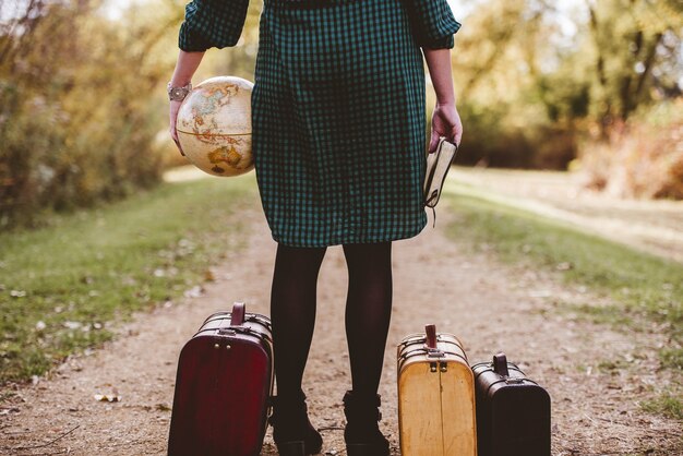 Женщина стоит на пустой дороге возле своего старого чемодана, держа Библию и настольный глобус