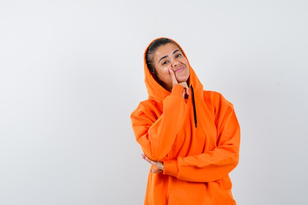 Female standing in thinking pose in orange hoodie and looking cheerful 