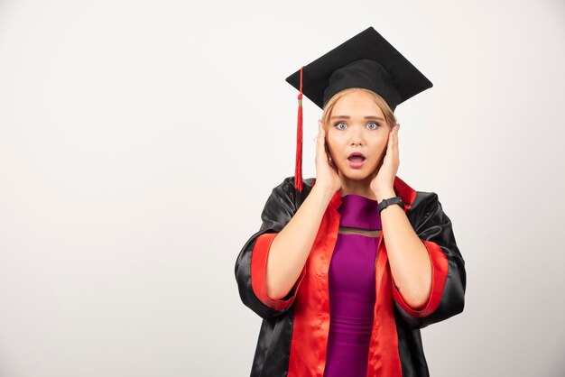 Female student in gown holding her face on white.