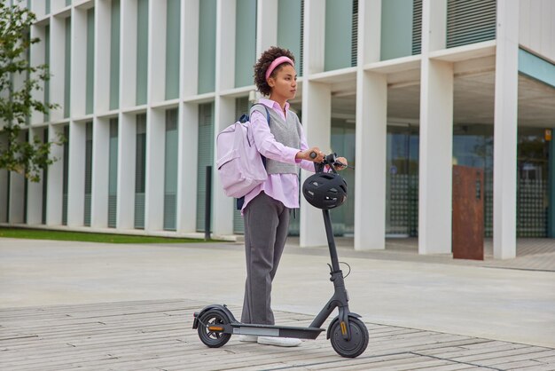 Female student rides to university by electric scooter carries rucksack poses against city buildings outdoors wears eleagant clothes