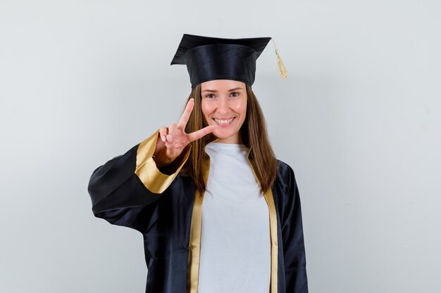 Студентка показывает V-знак в выпускном платье и выглядит веселым, вид спереди.