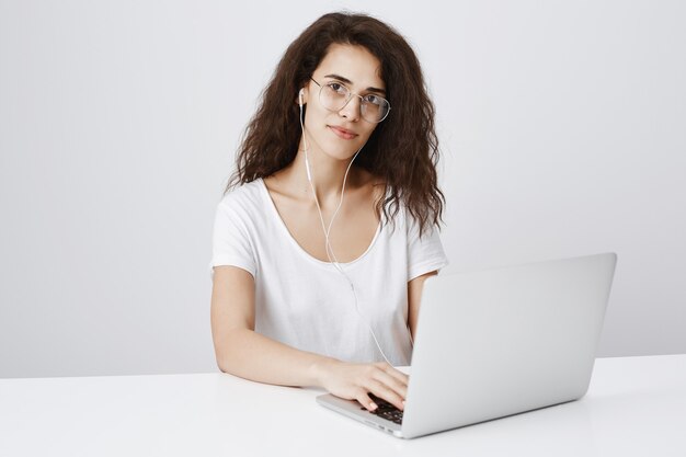 Female student sitting desk, using laptop and listening music in earphones