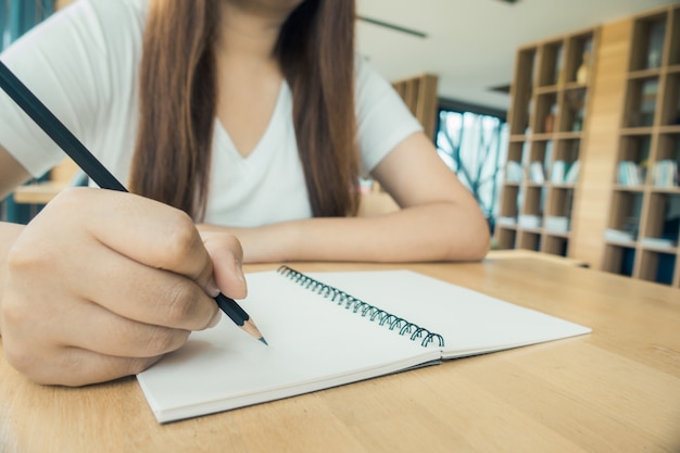 女性の学生が図書館の本からメモを取っています。若いアジア人の女性は、大学の図書館で課題を行うテーブルに座っています。ヴィンテージエフェクトスタイルの写真。