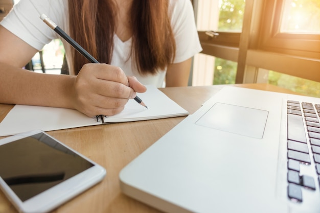 女性の学生が図書館の本からメモを取っています。若いアジア人の女性は、大学の図書館で課題を行うテーブルに座っています。ヴィンテージエフェクトスタイルの写真。