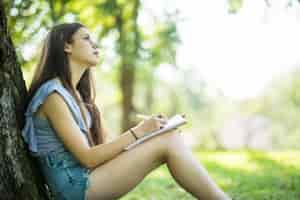 Free photo female student taking notes in her notebook. young smiling woman sitting in the park doing assignments. campus life, education, inspiration concept