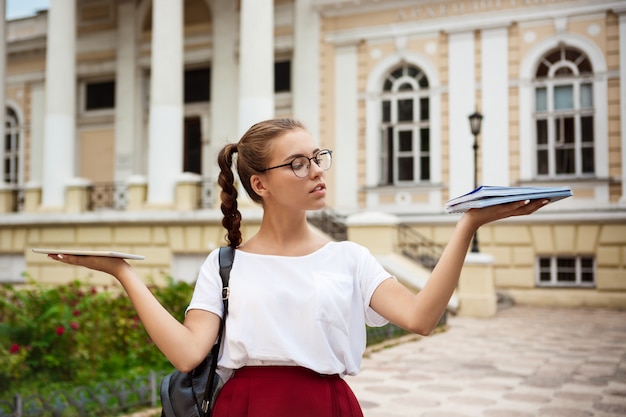 Female student wearing glasses holding tablet and notebooks in different hands outdoors.