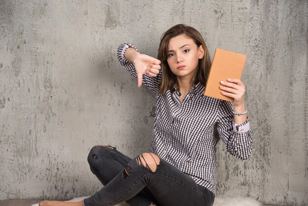 親指を下に与えるオレンジ色の本を持つ女子学生。