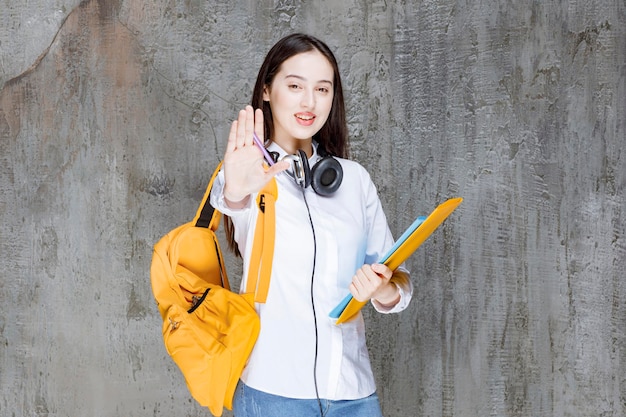노란색 배낭과 헤드폰을 들고 책을 들고 서 있는 여학생. 고품질 사진
