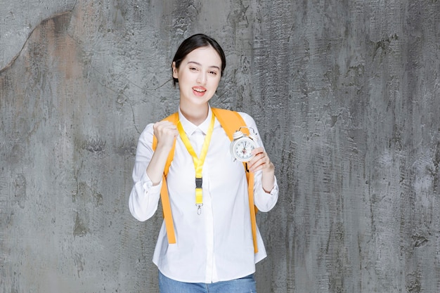 Female student with yellow backpack holding clock. High quality photo