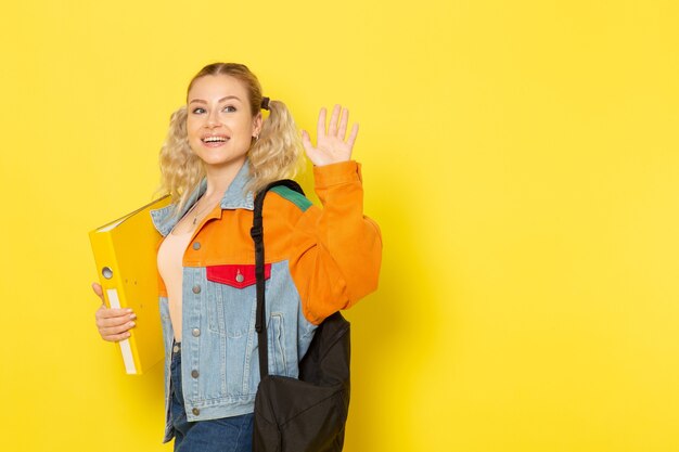female student young in modern clothes just simply posing with smile holding file waving on yellow