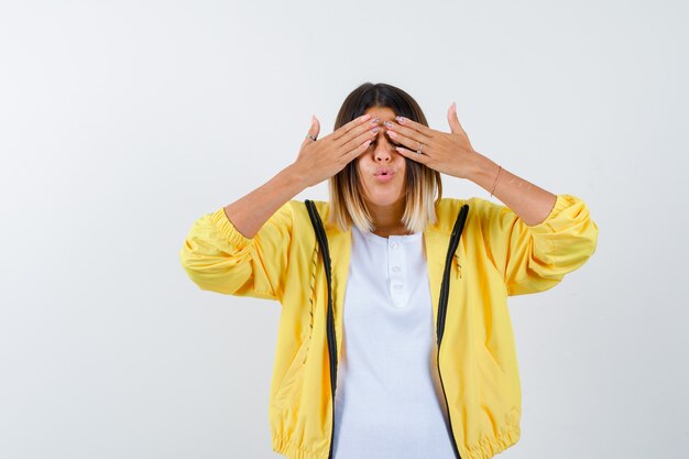 Female in t-shirt, jacket keeping hands on eyes, pouting lips and looking excited , front view.