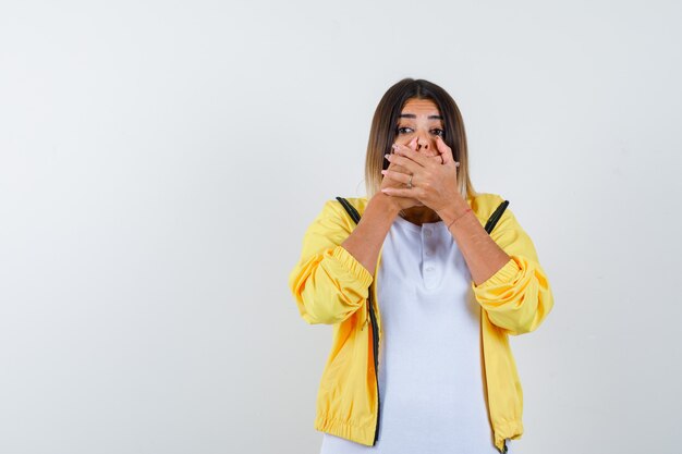 Female in t-shirt, jacket keeping hands on mouth and looking scared , front view.