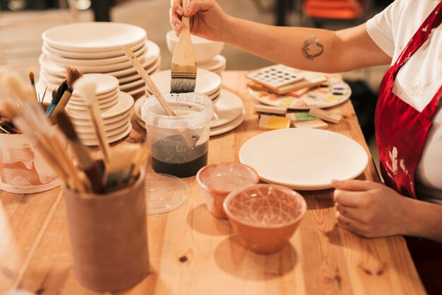 Женщина берет керамический цвет, чтобы рисовать на тарелке кистью