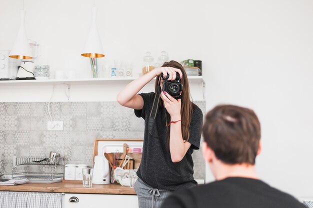 Female taking photograph of a man through camera in kitchen