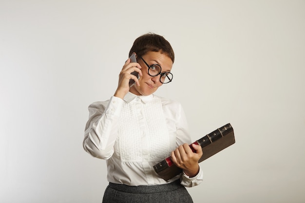 Учительница в консервативной одежде и круглых черных очках игриво смотрит по телефону на белой стене
