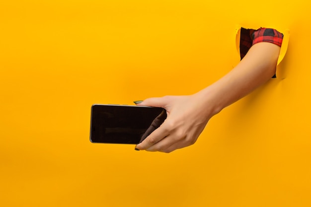 Female teen hands using mobile phone with black screen, through a torn yellow paper, isolated.