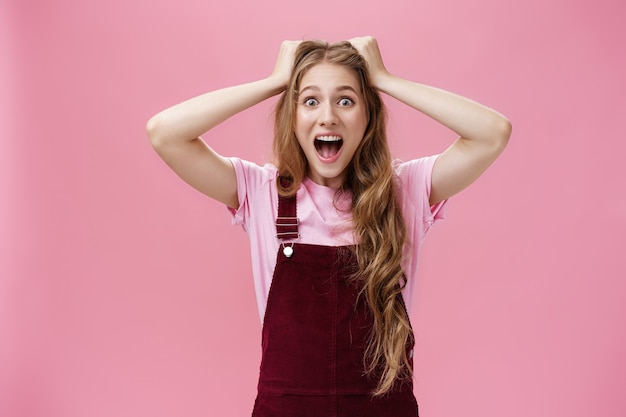 Female teenager cannot calm down feeling excited going on favorite band concert holding hands on head screaming from amazement and surprise standing overreacting against pink background.