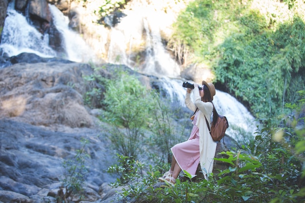 Female tourist who is looking at the binoculars to see the atmosphere at the waterfall