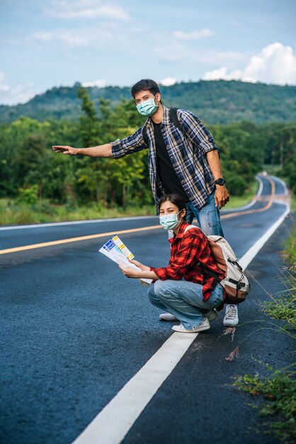 Female tourists sit and look at the map, male tourists pretending to hitchhike. Both wear masks and are on the side of the road.