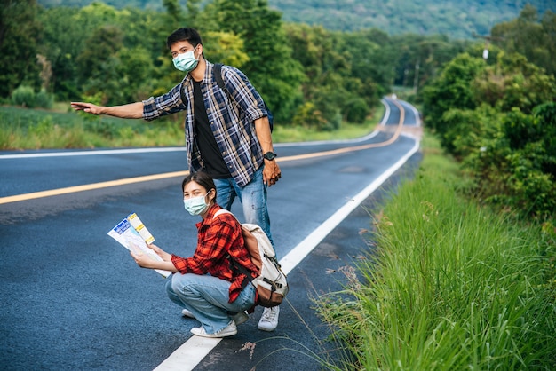 Female tourists sit and look at the map, male tourists pretending to hitchhike. Both wear masks and are on the side of the road.