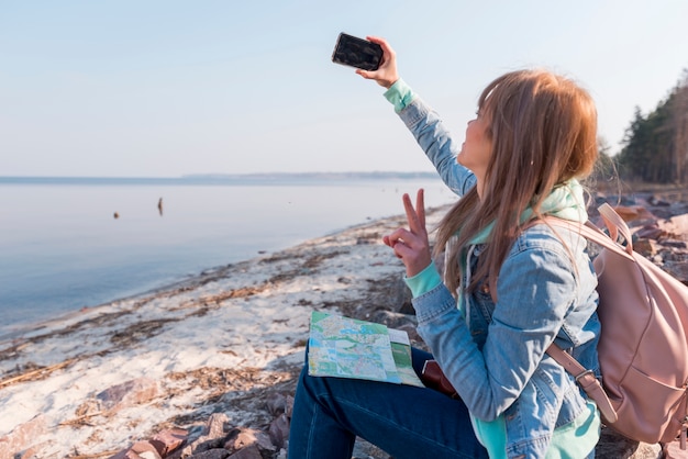 Женский путешественник, сидя на пляже, принимая селфи на мобильный телефон