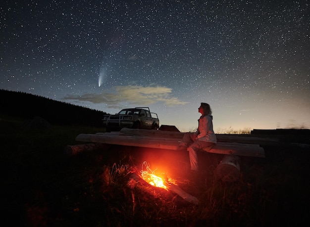 Female traveler sitting near campfire under night starry sky