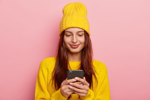 Female uses a smartphone, has long straight hair, wears yellow sweater and hat, has satisfied facial expression, poses against pink background