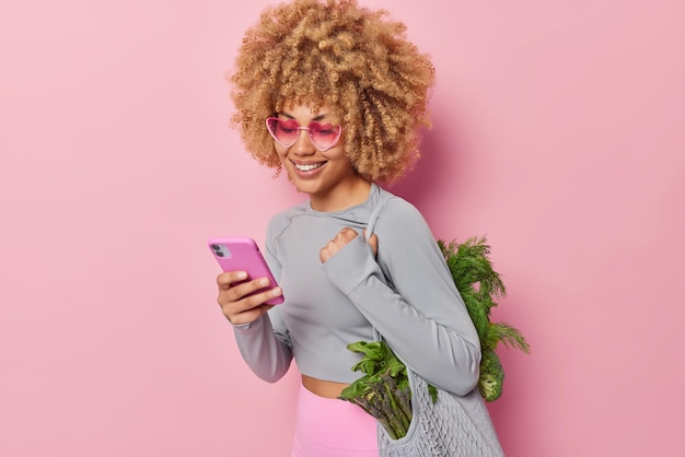 Женщина-вегетарианка с кудрявыми волосами носит сумку из органической ткани со свежими зелеными овощами, использует смартфон, просматривает меню, придерживается диеты, носит сердечки и спортивную одежду, изолированную на розовом фоне