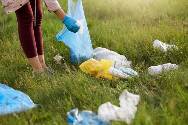 Female volunteer wearing leggins and gloves picking up litter in meadow, using blue garbage bag