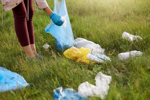 Женщина-волонтер в леггинсах и перчатках собирает мусор на лугу, используя синий мешок для мусора
