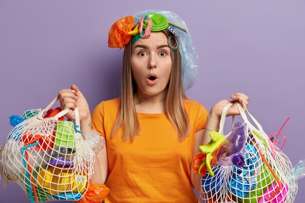 Female volunteer with astonished facial expression, collects garbage, holds two net bags, wears orange t shirt, can not believe she cleaned whole territory, stands against purple wall, recycles trash