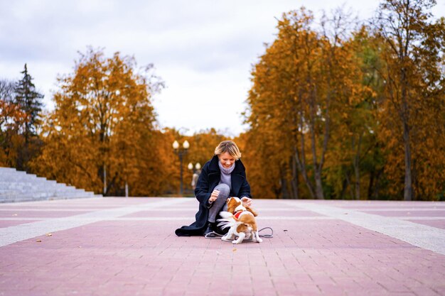 한 여성이 Cavalier King Charles Spaniel과 함께 공원을 걷고 있습니다. 강아지와 함께 가을 공원에서 산책 하는 여자. 카발리에 킹 찰스 스파니엘