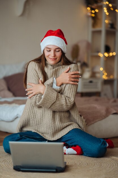 自宅でクリスマスのお祝いの間にラップトップでオンラインの友人と話している間笑顔のサンタを着ている女性