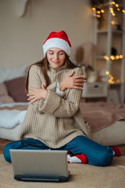 집에서 크리스마스 축하 기간 동안 노트북에 온라인 친구와 이야기하는 동안 웃는 산타를 입고 여성