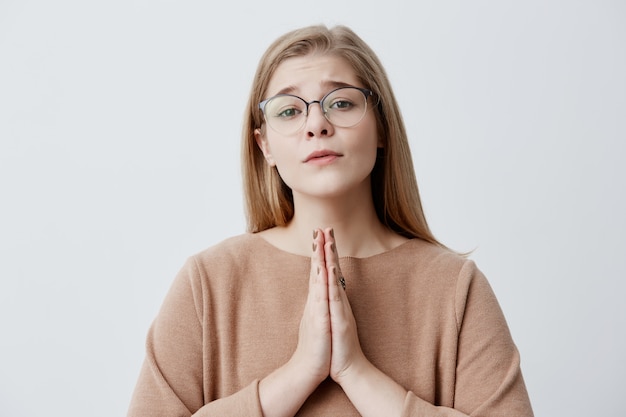 Female with dark eyes wearing eyeglasses having hopeful expression keeping her hands together asking for something hoping. Natural blonde female model anticipating for something important in life