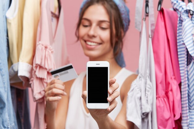 Female with happy expression standing near rack with clothes, keeping in hands credit card and modern mobile phone, being glad to buy garment online. People, online shopping