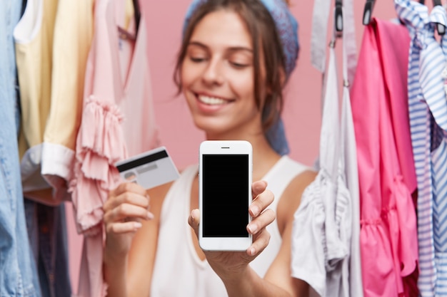 행복 한 표정으로 옷 선반 근처에 서, 손에 신용 카드와 현대 휴대 전화를 유지, 온라인 의류를 구입 기쁘다. 사람, 온라인 쇼핑