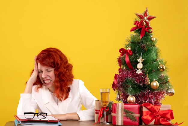 female worker sitting behind table with christmas presents and tree feeling angry on yellow
