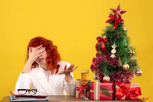 female worker sitting behind table with christmas presents and tree stressed on yellow