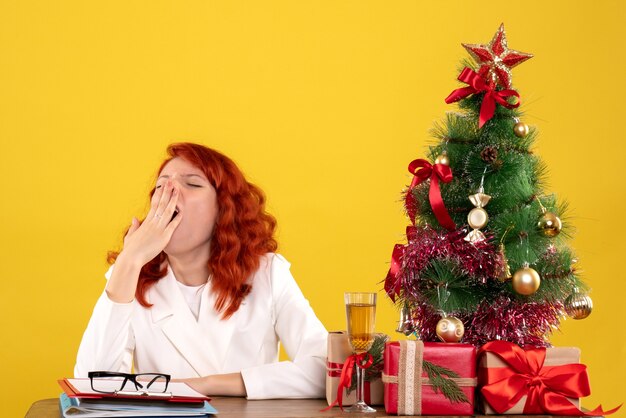 female worker sitting behind table with christmas presents and tree yawning on yellow