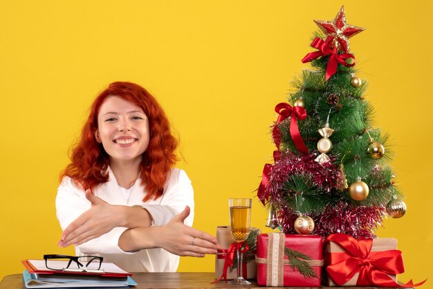 female worker sitting behind table with christmas presents and tree on yellow floor office xmas new year color