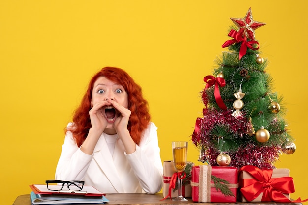 female worker sitting behind table with christmas presents and tree on yellow