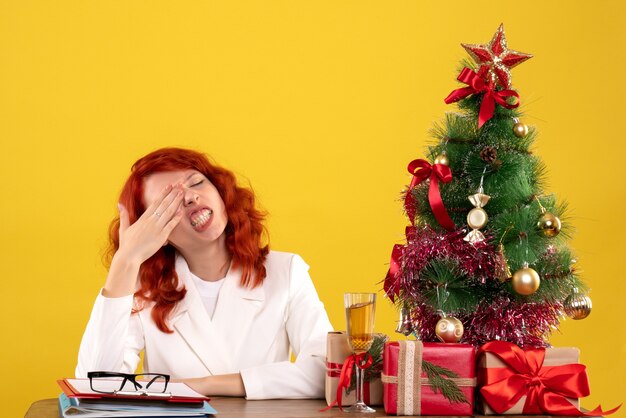 female worker sitting behind table with christmas presents and tree on yellow