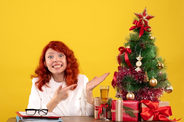 female worker sitting behind table with christmas presents and tree on yellow
